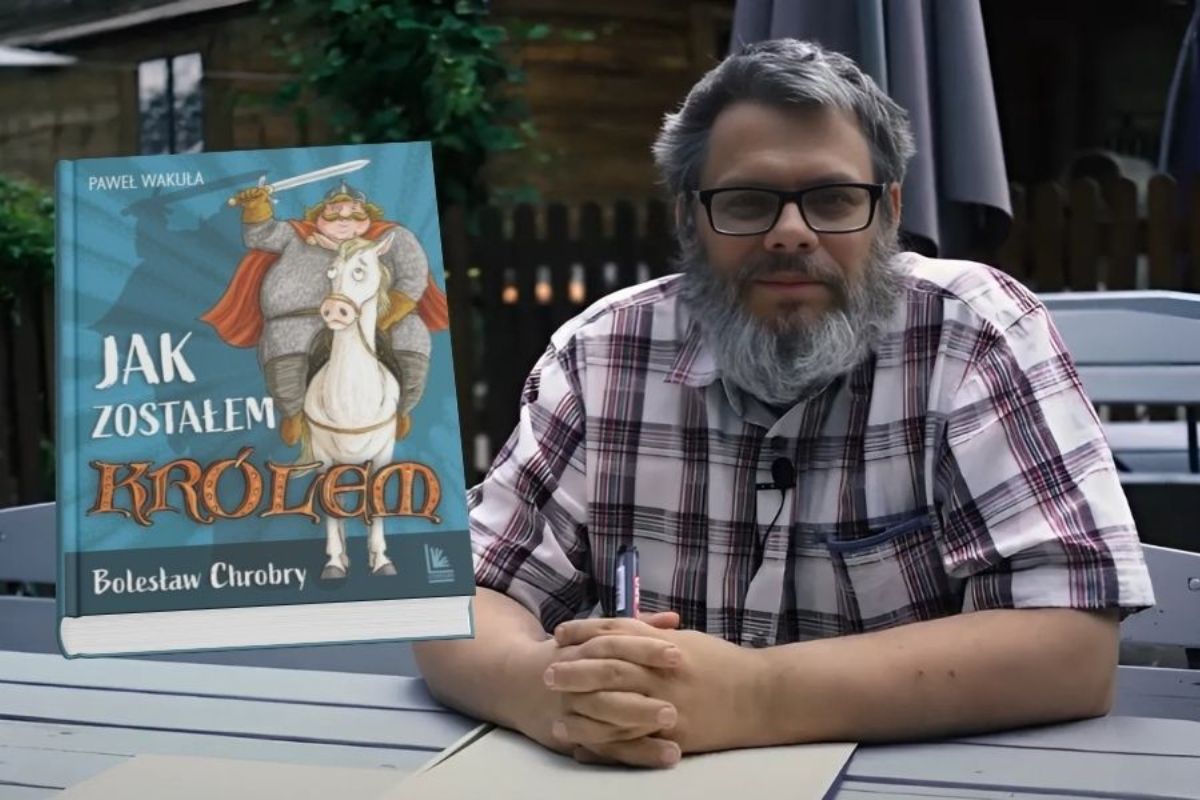 Mężczyzna z brodą siedzi przy stole, trzyma długopis, po lewej wmontowana okładka książki