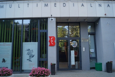 Fragment frontu biblioteki. Na górze na fasadzie napis: Multimedialna. Poniżej z lewej duże okno z dwoma plakatami. Po środku na słupku logotyp bibliotek warszawskich. Po prawej nowoczesne, szklane drzwi wejściowe.