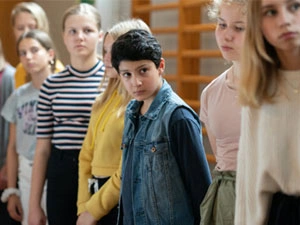 Grupa nastolatek i jeden chłopiec, Rami, stoją pod ścianą sali gimnastycznej i patrzą na lewo.