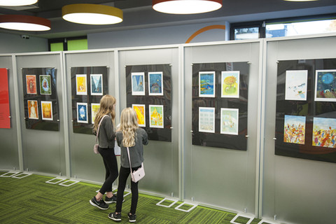 Dwie dziewczynki stoją przed planszami wystawienniczymi, wpatrują się w kolorowe szkice i ilustracje do książki „Aliaszka”. W tle widać salę biblioteczną.