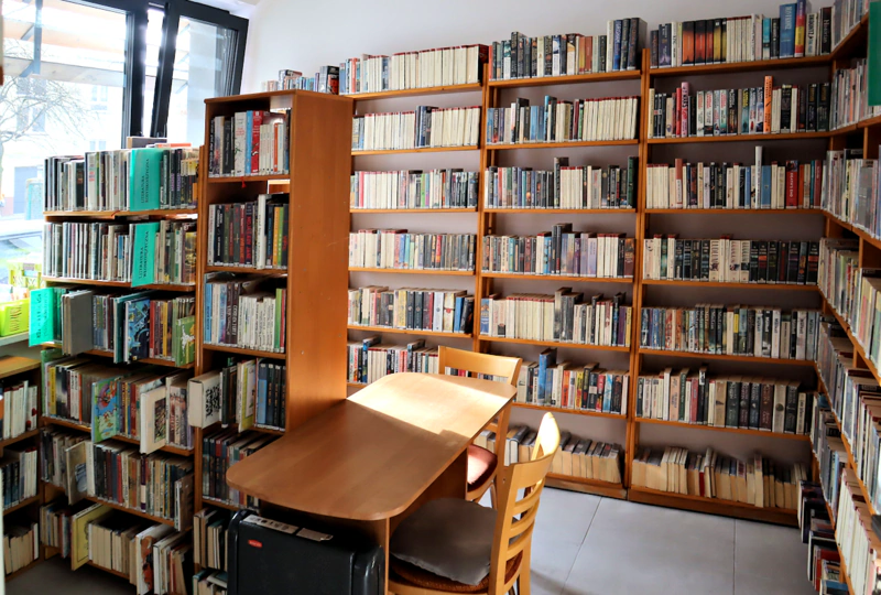 Okno z lewej. Na wprost i prawej wysokie, brązowe półki pełne książek. Pośrodku drewniany stolik z dwoma drewnianymi krzesłami. Przy oknie, obok stolika niższe drewniane regały z książkami.
