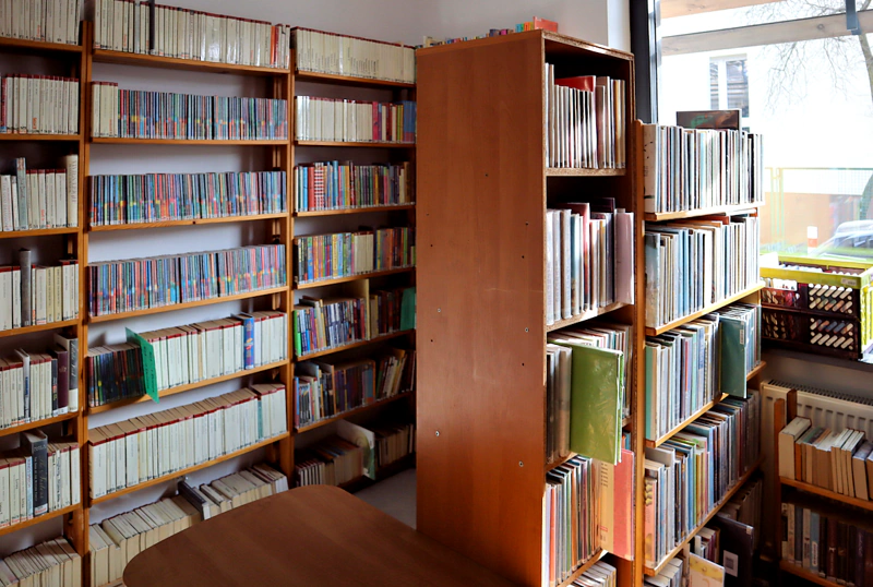 Z lewej przy ścianie wysoki drewniany regał pełny książek. Pośrodku przytulony do okna drewniany nieduży regał z książkami.