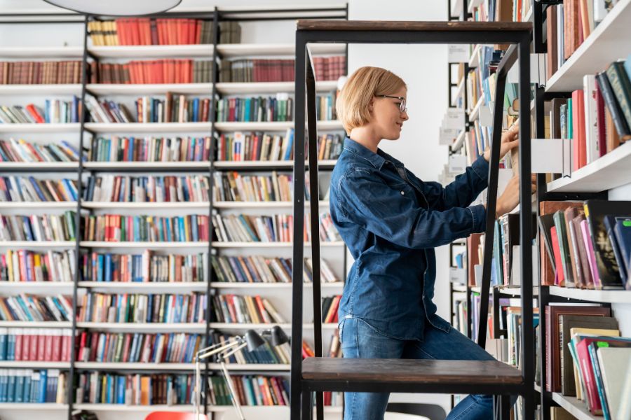 Kobieta stoi na schodkach w bibliotece i układa książki