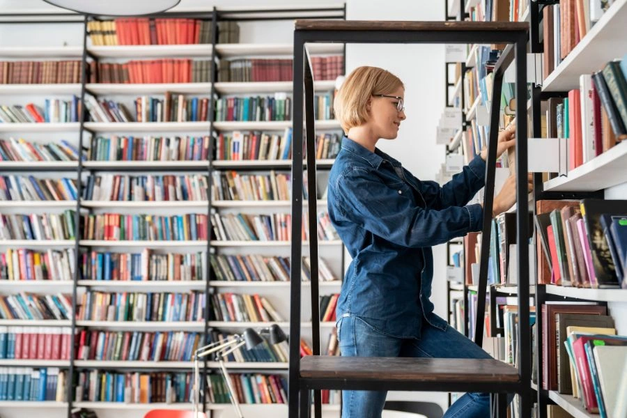 Kobieta stoi na schodkach w bibliotece i układa książki