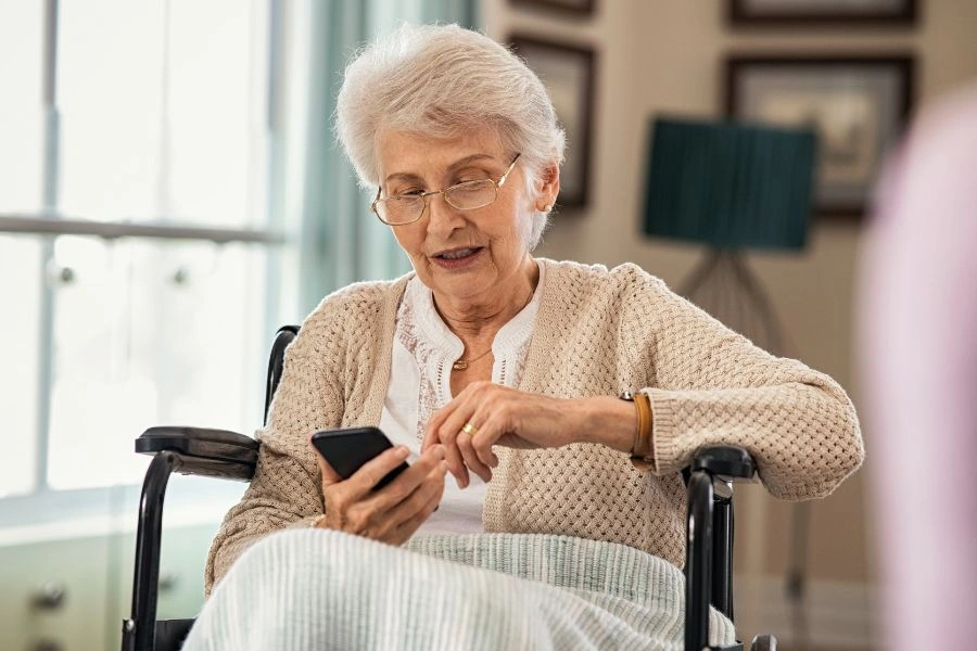Starsza kobieta siedzi na wózku inwalidzkim, trzyma w ręku smartfon