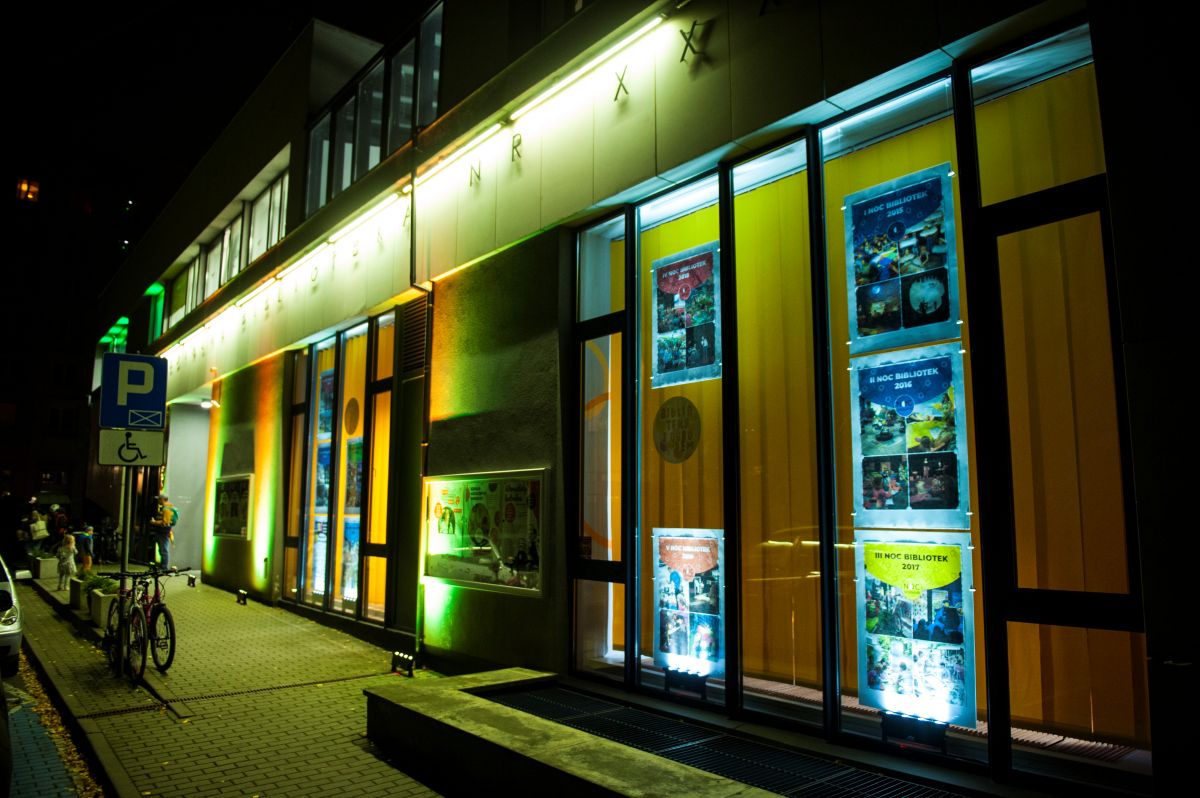 Nocna sceneria. Budynek Multimedialnej Biblioteki dla Dzieci i Młodzieży oświetlony światłem punktowym. W oknach widoczne kolorowe plakaty.
