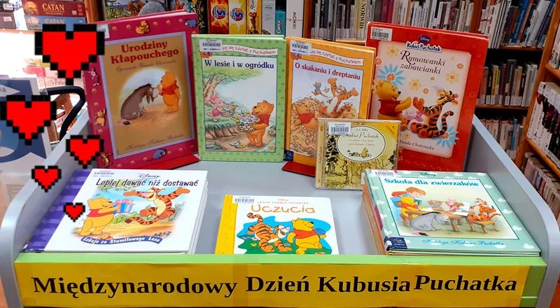 Stojak z na książki, na którym zostały umieszczone pozycje związane z Kubusiem Puchatkiem. Pod spodem tekst: międzynarodowy dzień Kubusia Puchatka. Z lewej kilka dorysowanych serduszek. W tle półka z książkami.