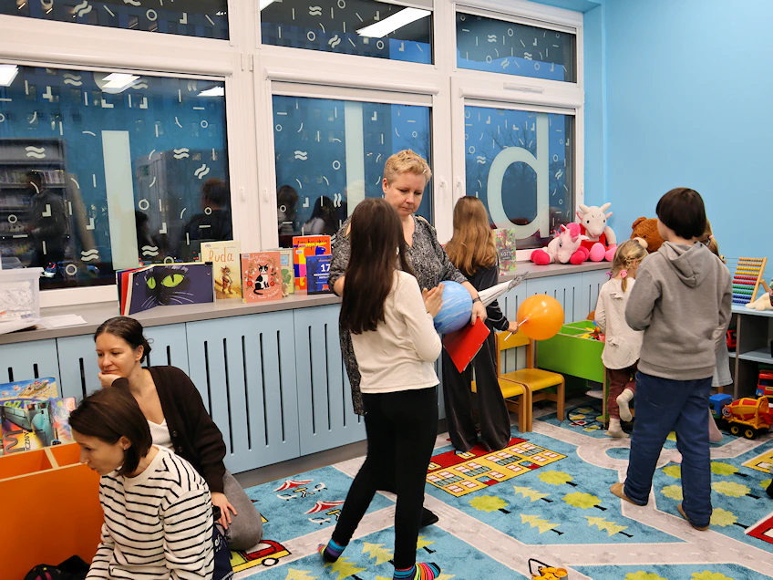 Bibliotekarka rozmawia z dziewczynką. Troje innych dzieci po prawej przygląda się zabawkom. Po lewej widać dwie kucające kobiety. Pod wszystkimi znajduje się wykładzina obrazkowa.