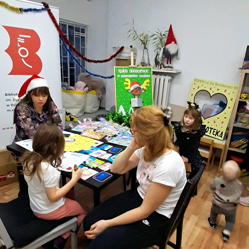 Przy czarnym stole ciemnowłosa bibliotekarka z czapką Mikołaja na głowie rozmawia z ciemnowłosą dziewczynką rysującą coś na kartce. Przy stole siedzi jeszcze inna dziewczynka i jasnowłosa nastolatka. Obok nich spaceruje około dwuletnie dziecko.