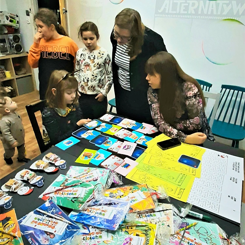 Dziewczynka z siedzi przy czarnym stole odgadując zagadkę świąteczną. Kibicuje jej dwójka dorosłych kobiet. Obok stoją jeszcze dwie inne dziewczynki. Patrzy na nie stojące obok około dwuletnie dziecko.