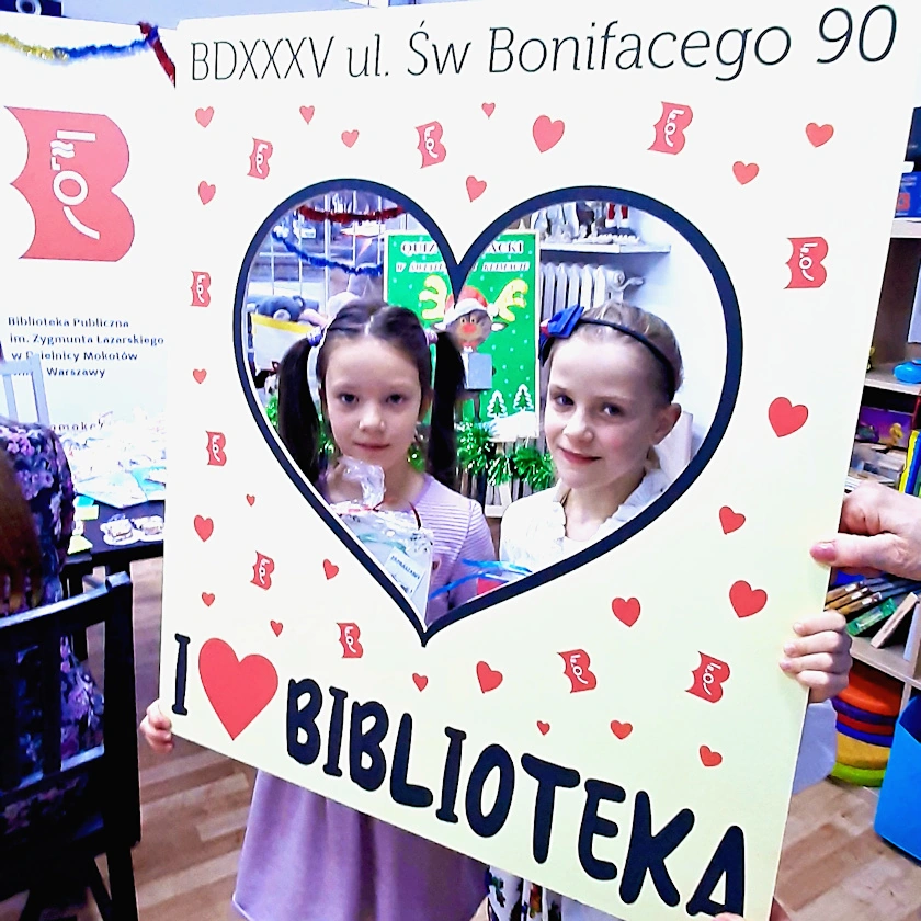 Fotoramka z napisem u górze b d 35 ul. Świętego Bonifacego 90. Na dole napis Aj love biblioteka. Pośrodku wycięty otwór w kształcie serca, a w nim widać dwie uśmiechnięte dziewczynki.
