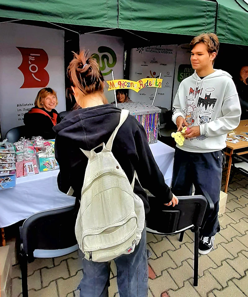 Dziewczyna i chłopak w wieku licealnym stoją przy stoisku mokotowskiej biblioteki. Bibliotekarka siedzi na krześle i uśmiecha się do nich. Na stole leżą książeczki dla dzieci.