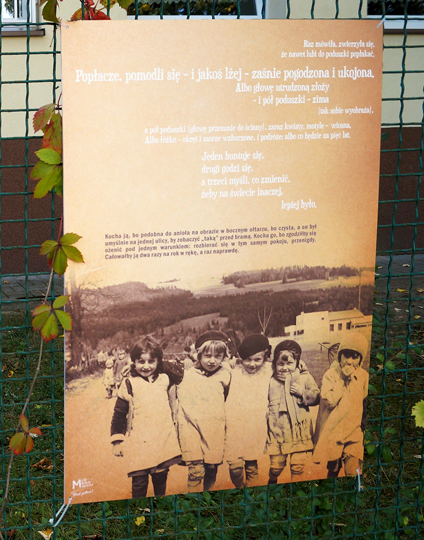 Plansza powieszona na drucianym, zielonym płocie. Na górze planszy jakiś tekst. Poniżej zdjęcie idących gdzieś małoletnich dziewczynek. W ich tle niski budynek i zalesione wzgórze.
