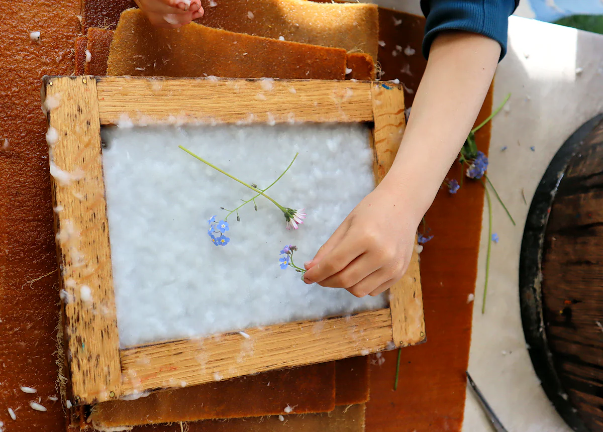 sitko osadzone na drewnianej ramie a w nim susząca się papierowa pulpa. Ręka młodej osoby dekoruje pulpę niebieskimi kwiatkami.