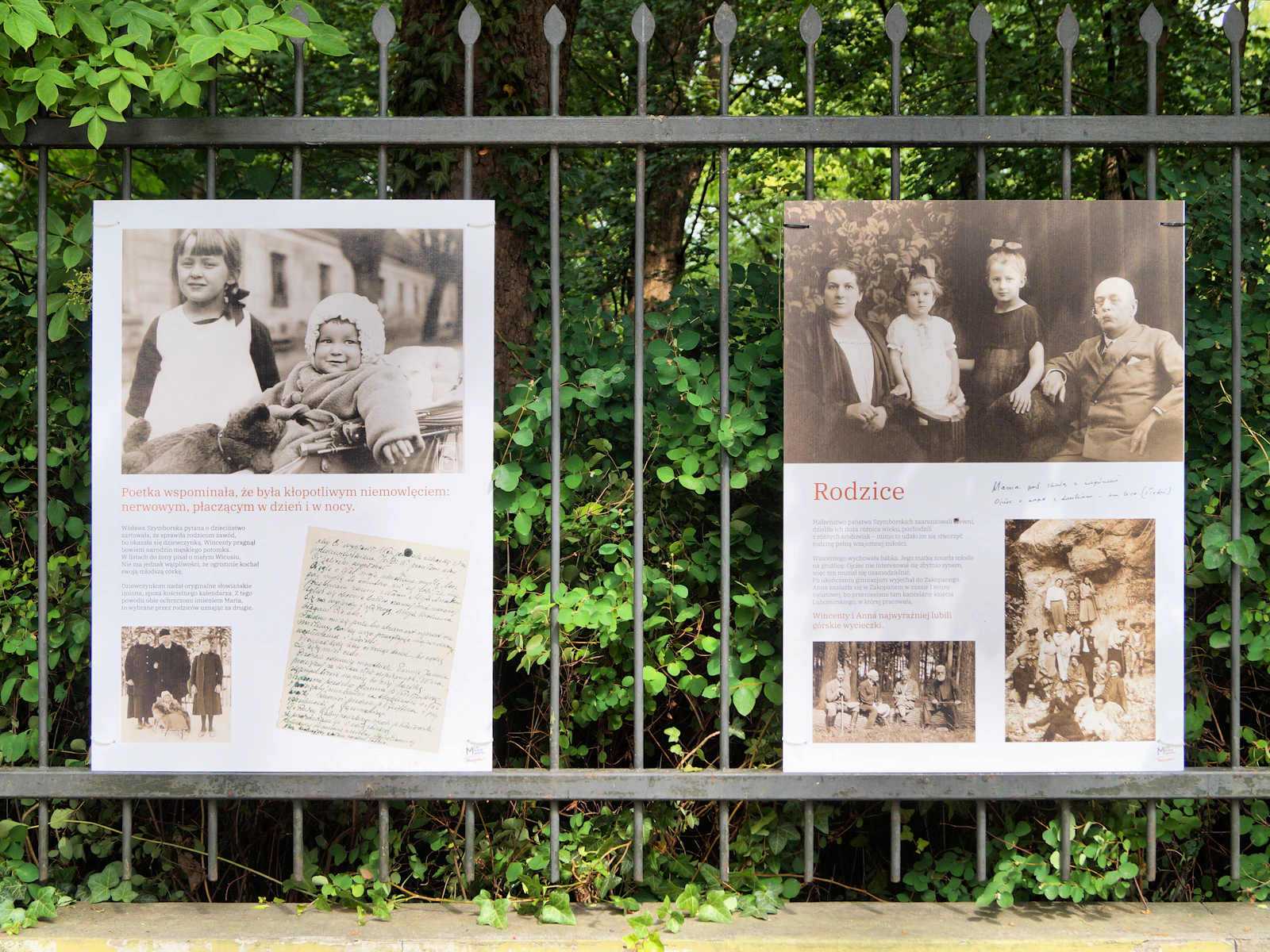 W tle gęsto od zieleni. Na żelaznym ogrodzeniu powieszane dwie duże plansze. Obie z różnymi zdjęciami z dzieciństwa Szymborskiej z jakimś dodatkowym tekstem.