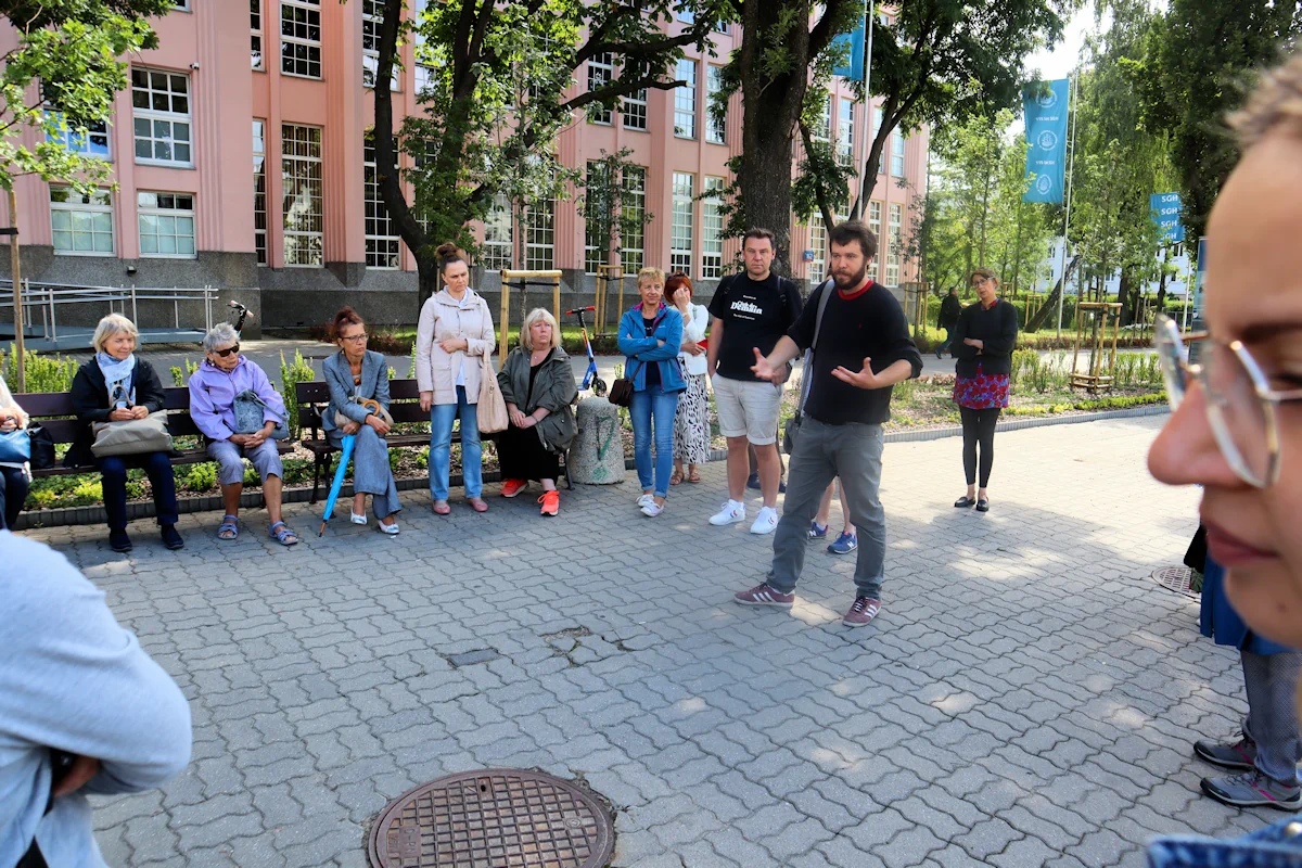 Grupa ludzi zgromadzona wokół przewodnika pod głównym budynkiem Wyższej Szkoły Handlowej. Pogoda dopisuje, jedni ubrani letnio, inni wiosennie.