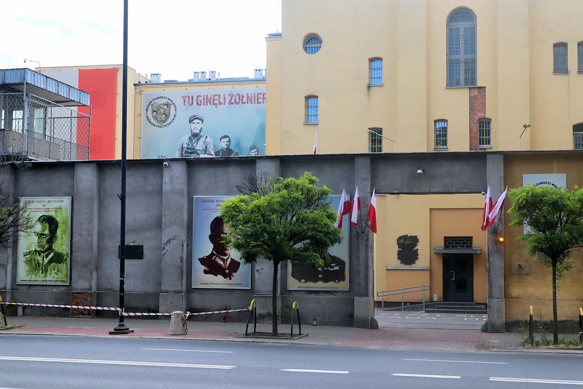 Brama w wysokim murze, a za nim budynek byłego aresztu śledczego, obecnie Muzeum Żołnierzy Wyklętych i więźniów politycznych. Mur jest szary, częściowo przesłonięty drzewami, a na nim portrety trzech więźniów. Przed bramą flagi Polski. Budynek ma wąskie okna, tynk w kolorze ciemno żółtym. Z lewej strony nad murem widać fragment wieży strażniczej.