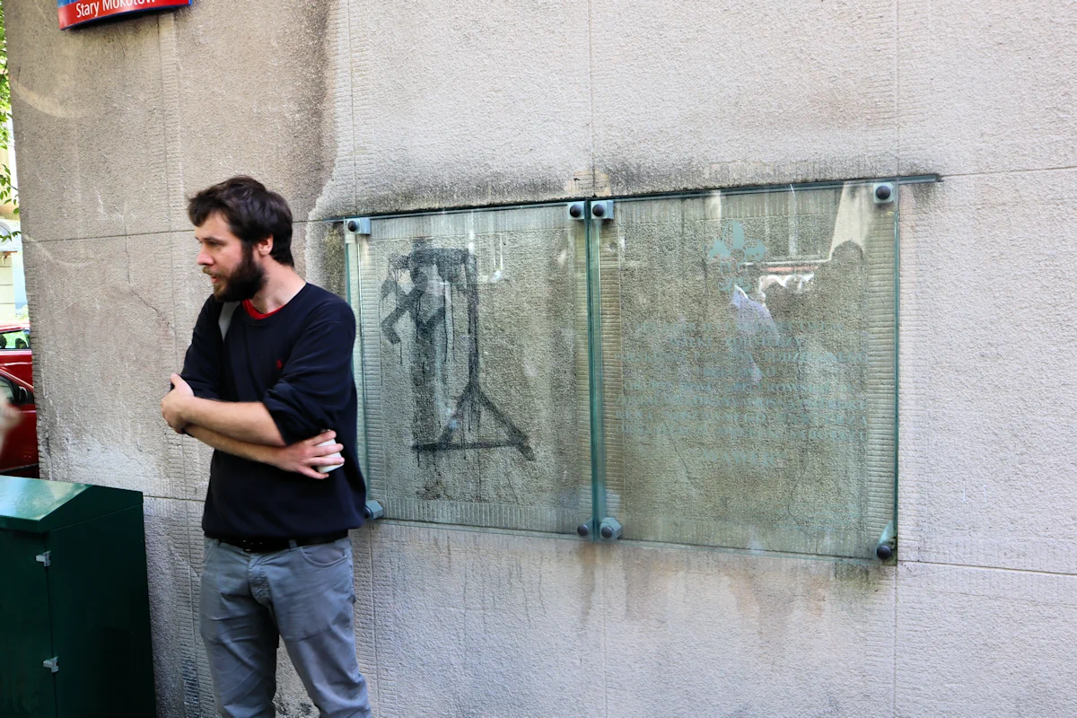 Przewodnik stoi obok zachowanego graffiti z czasów powstania warszawskiego. Grafika jest zasłonięta szkłem, przedstawia swastykę powieszoną na szubienicy. Obok tablica informacyjna niestety nie do odczytania.