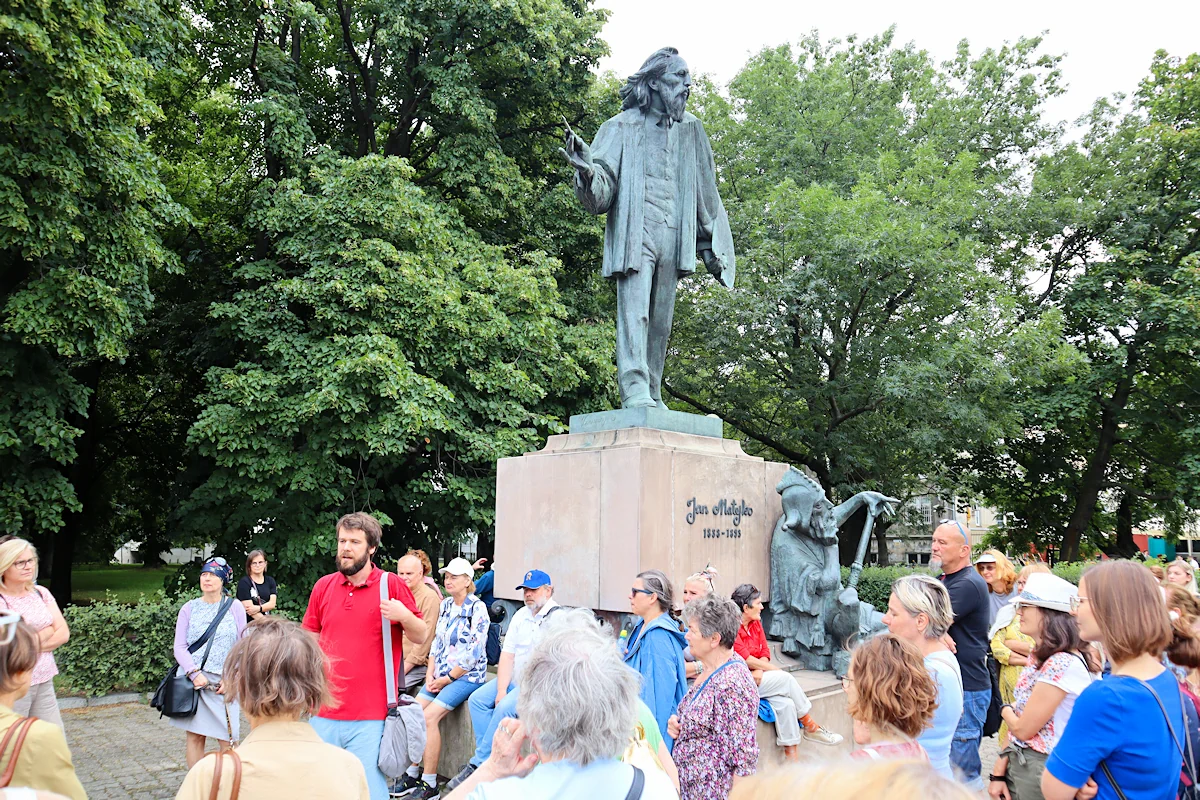 Grupa osób zgromadzonych przy pomniku Jana Matejki słucha słów przewodnika. Matejko w prawej ręce trzyma pędzel, w lewej paletę. U stóp cokołu widać rzeźbę siedzącego Stańczyka, zamyślonego, z buławą w lewej ręce. W tle rozpościera się gęsta zieleń.