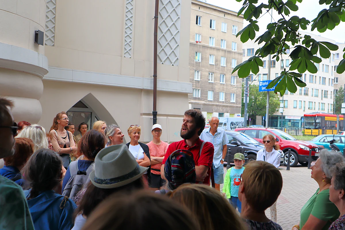 Grupa osób słucha przewodnika na jeden z ulic Mokotowa. Po lewej widać kawałek zabytkowego budynku. W głębi samochody, tramwaj i budynki po drugiej stronie ulicy. Kawałek gałęzi częściowo przesłania kadr z prawej strony.