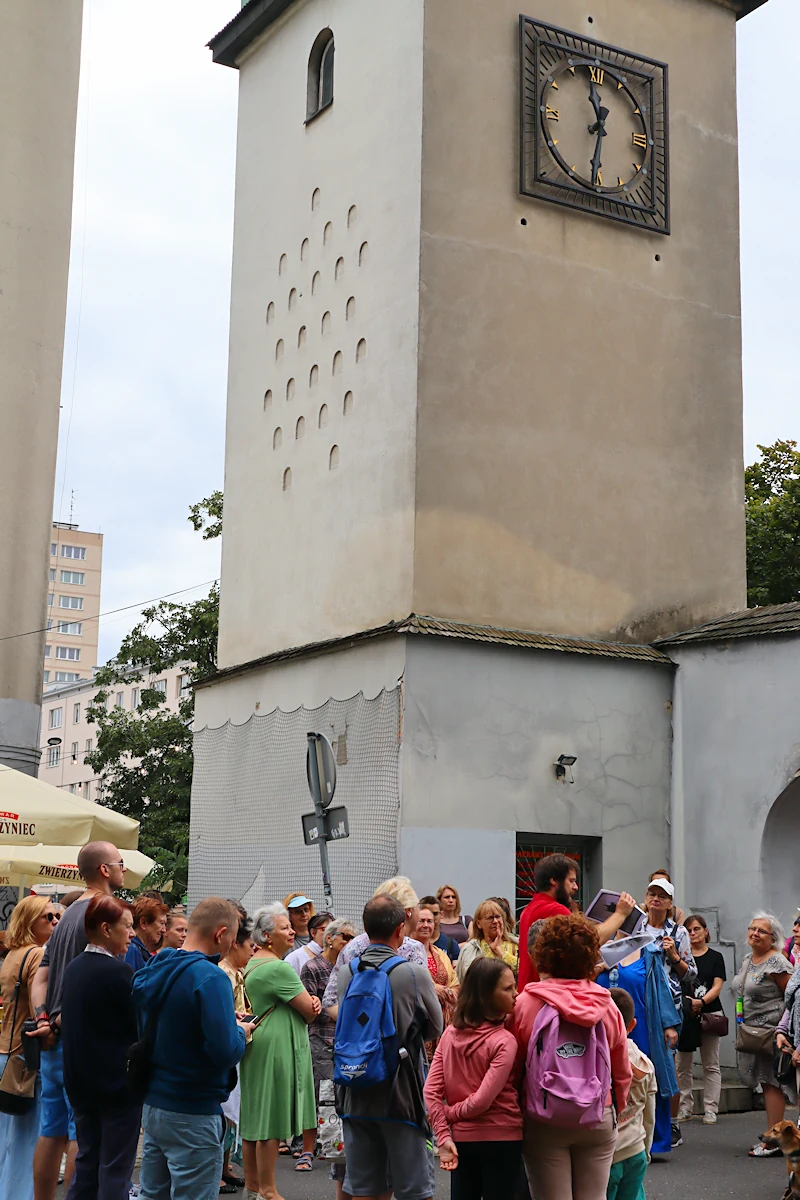 Kilkadziesiąt osób stoi przy starej budowli. Jest to fragment niezbyt dużej bramy i niewysoka wieża z zegarem mechanicznym. Wskazówki pokazują jedenastą trzydzieści. W tle widać zieleń i bardziej współczesne budynki.