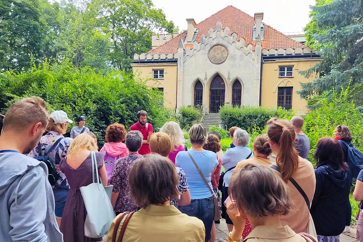 Grupa osób stoi tyłem, słuchając przewodnika, patrzy na historyczną budowlę. Jest to niewielki pałacyk dwupoziomowy. Wejście frontowe jest stylizowane na gotyk - wąskie strzeliste okna oraz wejście, wyglądają jak wejście do kościoła. Boki i dach to już raczej architektura klasycystyczna. Przed budynkiem dużo zieleni.