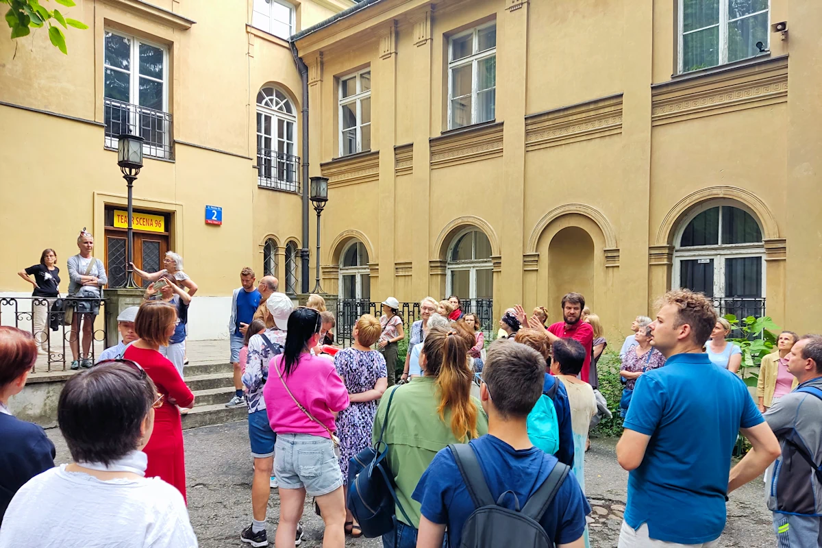 Grupa osób stoi na dziedzińcu historycznej budowli. Styl to chyba architektura klasycystyczna. Z prawej dwa poziomy z dużymi oknami. Z lewej prostopadle do niego trzypoziomowa konstrukcja z wejściem, nad którym widać napis: Teatr Scena 96. Do wejścia prowadzi nieduży taras, schodki i żelazne barierki. Przy schodach są dwie nieduże latarnie stylizowane na lampy gazowe.