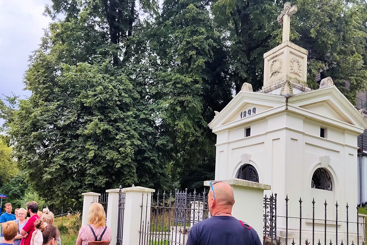 Z lewej widać kilka osób i przewodnika w czerwonej koszulce polo. Za obrodzeniem widać niewielki biały grobowiec w kształcie małej wieży, zwieńczony kamiennym krzyżem. Na frontowej ścianie cyfry: 1899. W tle widać kilka rosłych drzew.