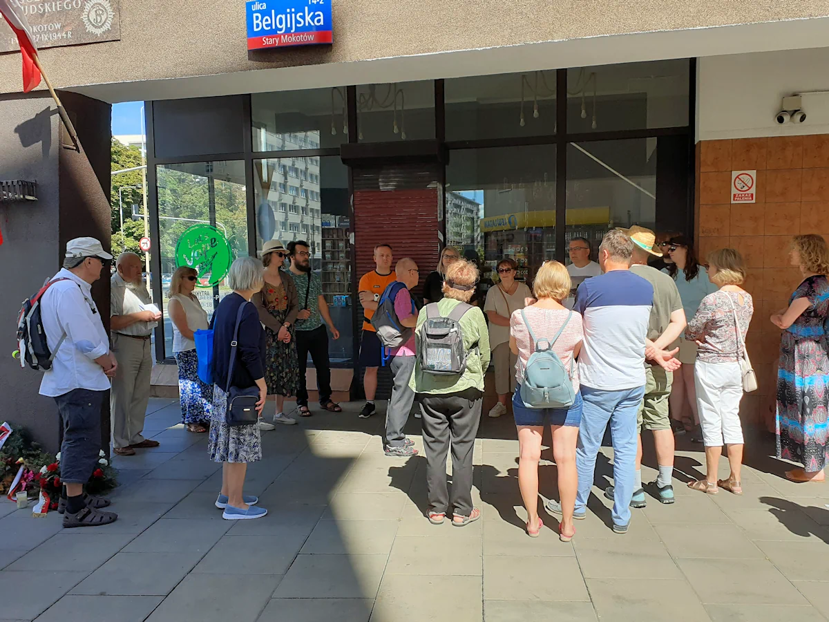 Ubrana letnio gromada osób zgromadzona na ulicy Belgijskiej. Budynek narożny z dużymi witrymani jakiegoś sklepu.