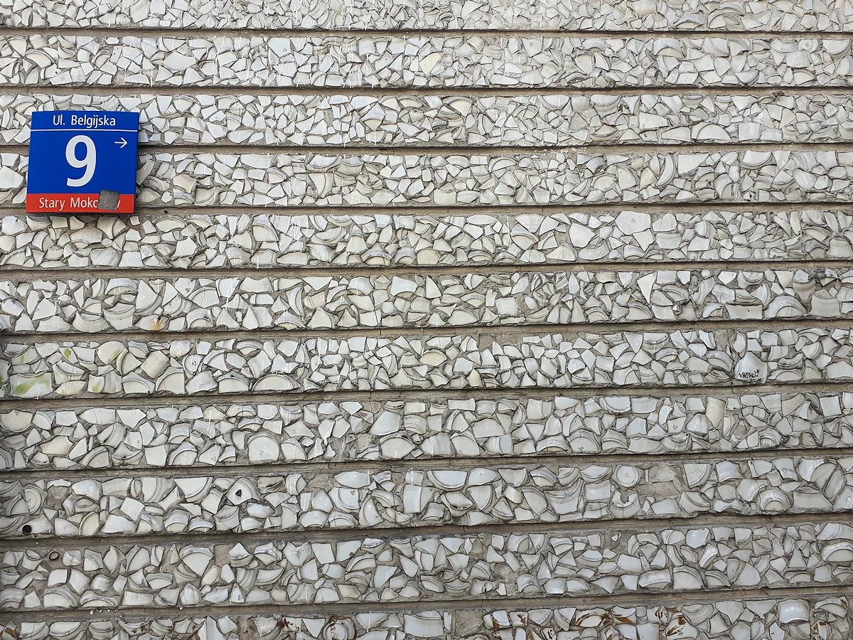 Ściana budynku wyłożona poziomymi pasami potłuczonych naczyń. W lewym górny rogu tablica informacyjna: ulica Belgijska 9; Stary Mokotów.