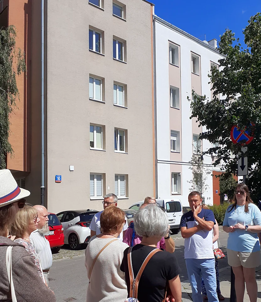 Gromada letnio ubranych osób stoi na tle trzypiętrowego budynku o szarych ścianach i przytulonego do niego budynku o białych ścianach. Z prawej fragment liściastego drzewa oraz znak zakaz zatrzymywania się.