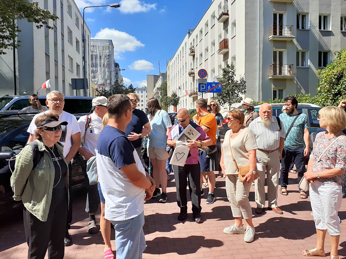 Gromada osób stoi na czerwonym chodniku. W tle ulica z kilkupiętrowymi budynkami po prawej i po lewej. Przewodnik coś chowa do swojej torby.