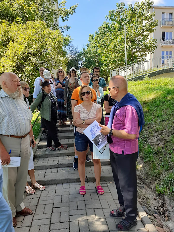 Gromada osób stoi na schodach i z uwagą słucha przewodnika, który trzyma w ręku szkice wcześniej tu obecnych budynków. W tle drzewa liściaste, a z prawej fragment kilkupiętrowego budynku.