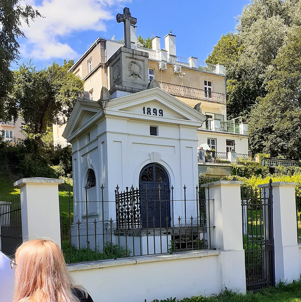 Rodzinna niewielka krypta w białej tonacji, z krzyżem na szczycie w szarej tonacji. W tle na wzniesieniu trzypoziomowy budynek z tarasem.