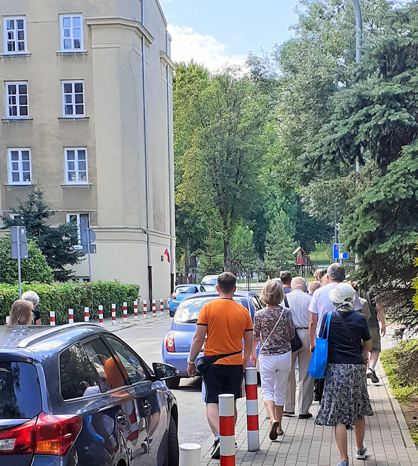 Idziemy za gromadą osób po chodniki z prawej strony ulicy. Stoją tam samochody. Z lewej fragment trzypiętrowego budynku o szaro-żółtych ścianach.