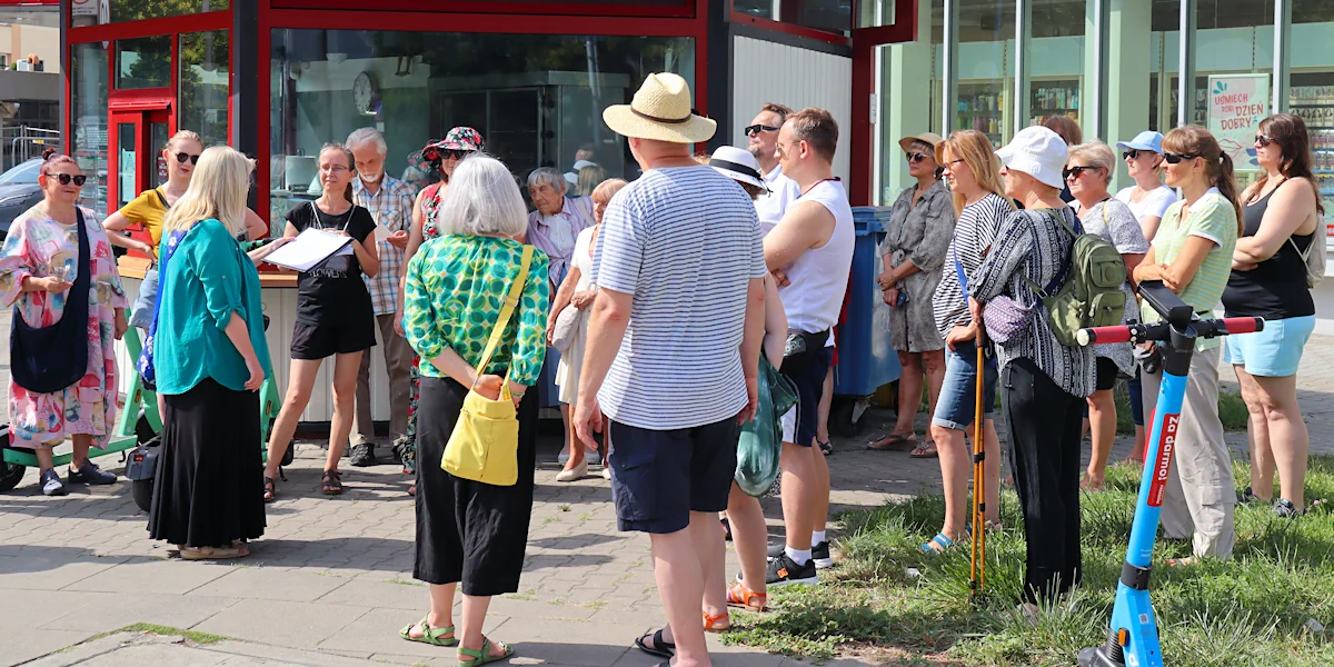 Grupa osób ubrana letnie zgromadziła się wokół przewodniczki ubraną w błękitną koszulkę z krótkimi rękawami i ciemnogranatową spódnicę. W tle szklane witryny kiosku z jedzeniem, a za nim inny budynek ze szklanymi witrynami.