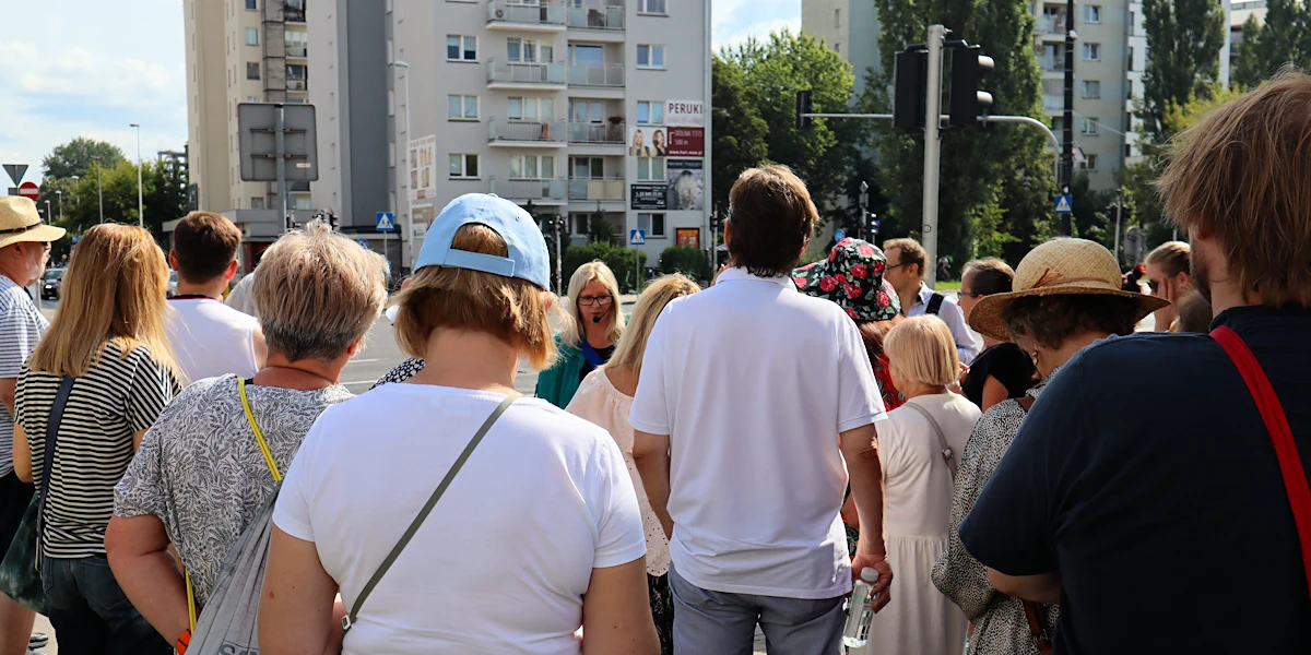 Plecy grupy ludzi stojących przy skrzyżowaniu. W tle widać słupy z sygnalizacją świetlną i dwa wieżowce oraz kilka wysokich drzew.