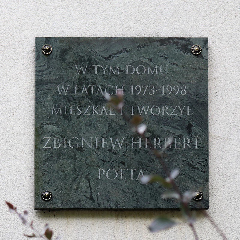 Na jasnej ścianie wisi tabliczka z mocno ciemnego marmuru. Na niej tekst: w tym domu w latach 1973 - 1998 mieszkał i tworzył Zbigniew Herbert, poeta.