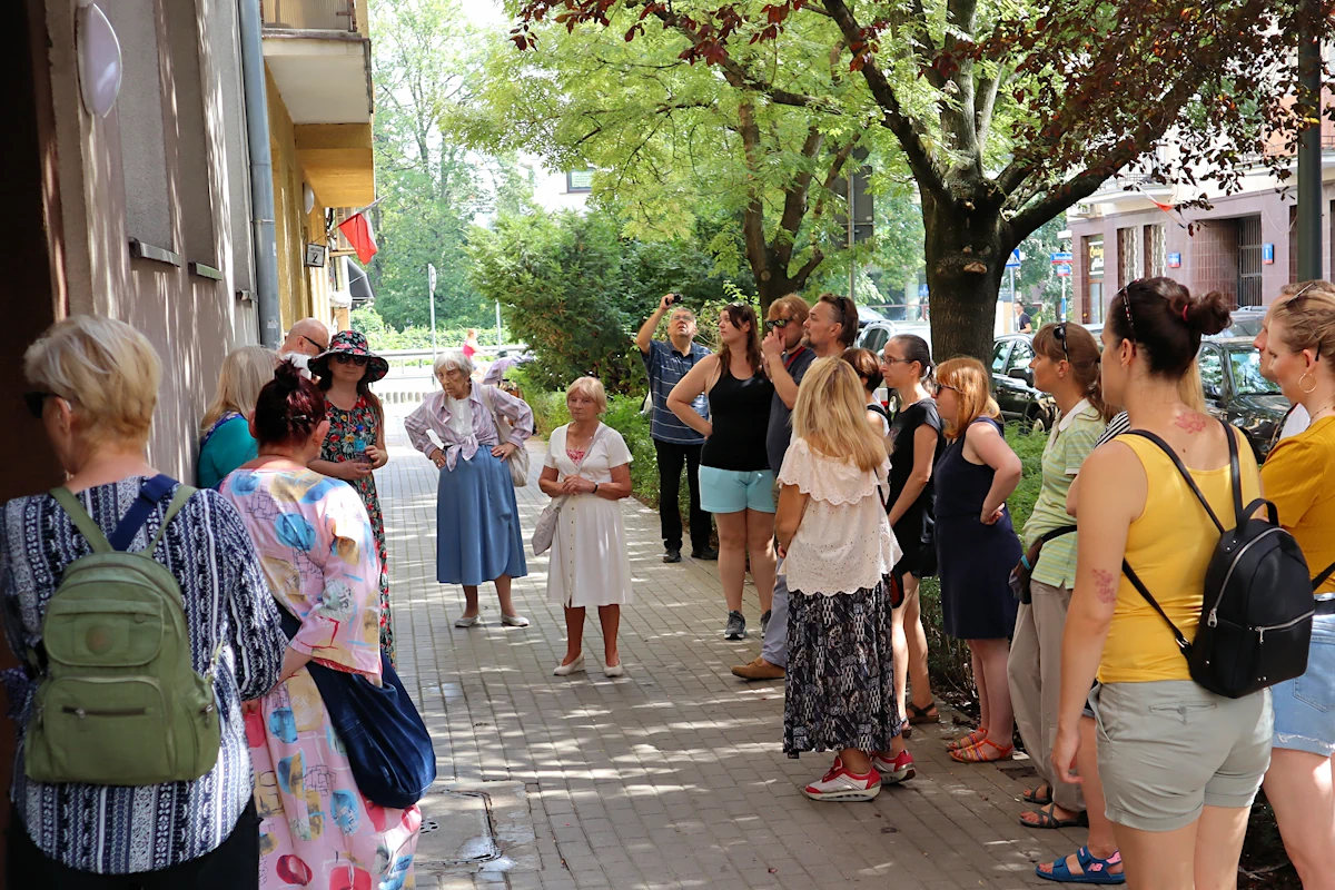 Grupa letnio ubranych osób stoi przed starym blokiem mieszkalnym, słuchając opowieści przewodniczki. Na ulicy są tez drzewa i krzaki.