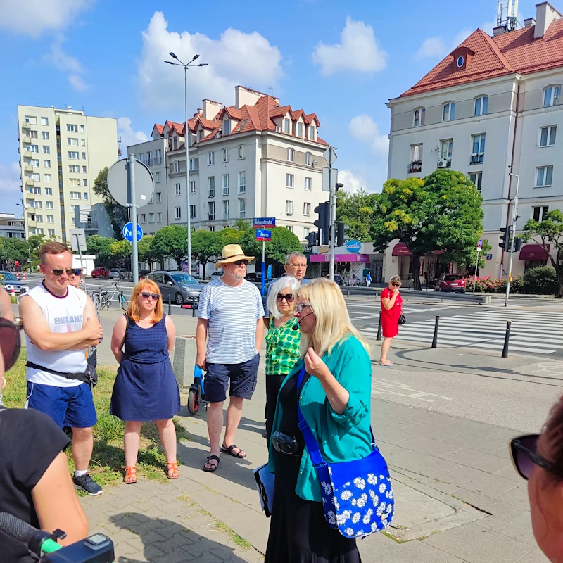 Grupa osób stoi na chodniku wokół przewodniczki. W tle widać ulicę kilkujezdną, na której widać dwa samochody osobowe. Po drugiej stronie ulicy widać kilka drzew, dwa czteropiętrowe domy i jeden wyższy budynek mieszkalny.