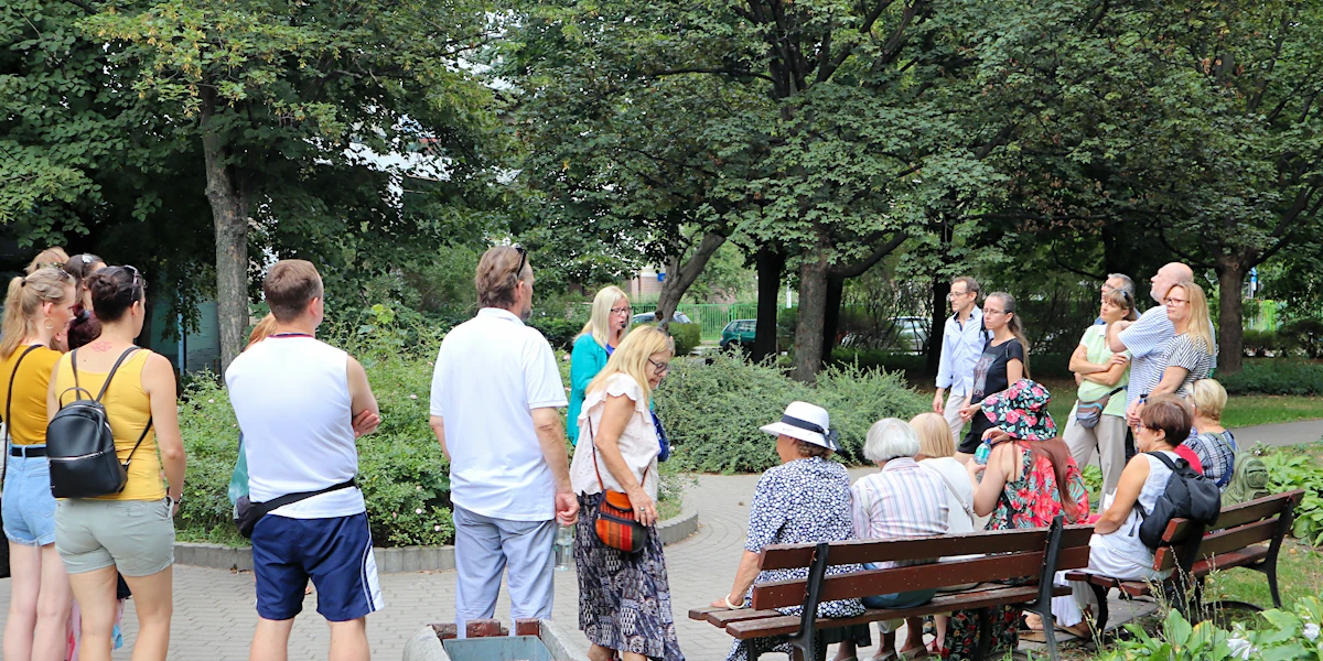 Część osób stoi, część siedzi w paru, wsłuchując się w opowieść przewodniczki. Poza alejką widać klomb z nisko rosnącą roślinnością. W tle wyrośnięte drzewa liściaste.