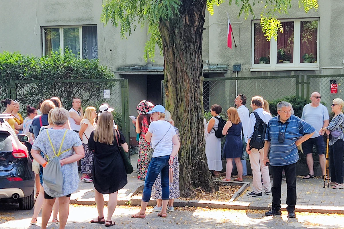 Grupa osób stoi przed zielonym ogrodzeniem, za którym widać front niewysokiego budynku. Budynek częściowo jest zasłonięty przez szeroki pień oraz wyrośnięte krzaki.
