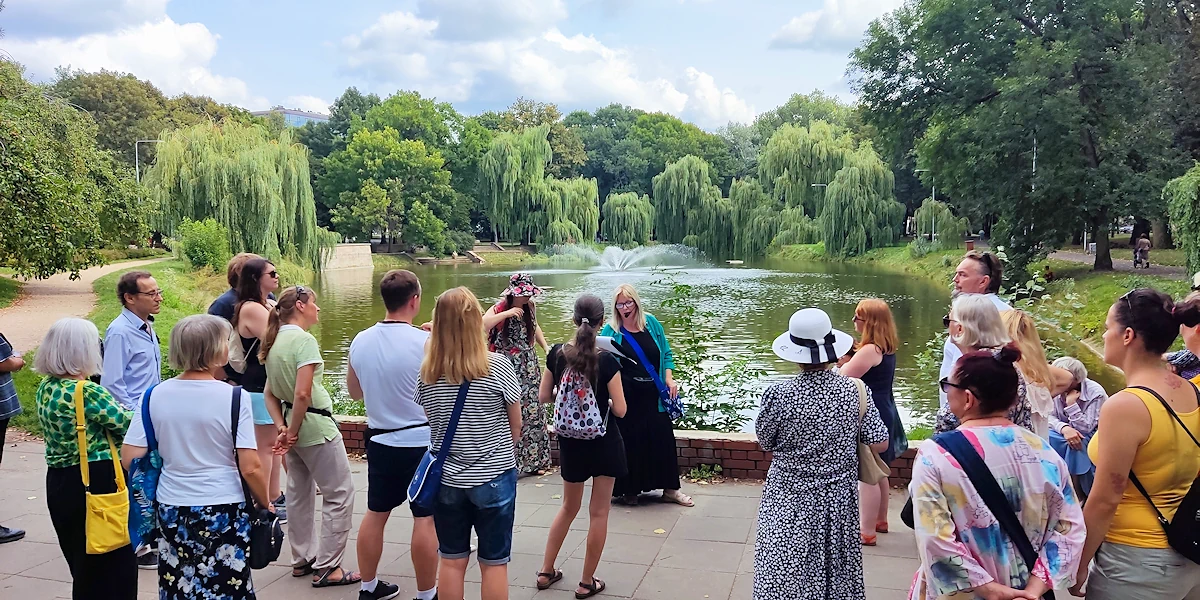 Grupa osób stoi na tle pięknego widoku soczyście zielonego parku z dużym zbiornikiem wodnym po środku. Ze środka zbiornika bije fontanna wody. Przewodniczka opowiada o czymś z ożywieniem.