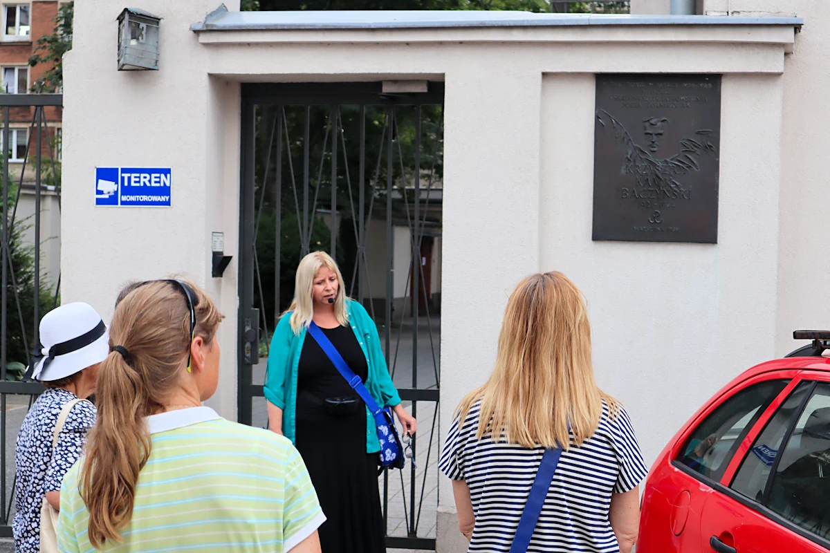 klika osób słucha przewodniczki. Stoją przed ogrodzeniem do budynku w głębi. Z prawej strony furtki, na białym tle muru wisi tabliczka poświęcona polskiemu poecie. Napis głosi: W tym domu w latach 1936 - 1944 mieszkał i stąd wyszedł do powstania poeta, żołnierz AK, Krzysztof Kamil Baczyński; 1921 - 1944; Warszawa 1989.