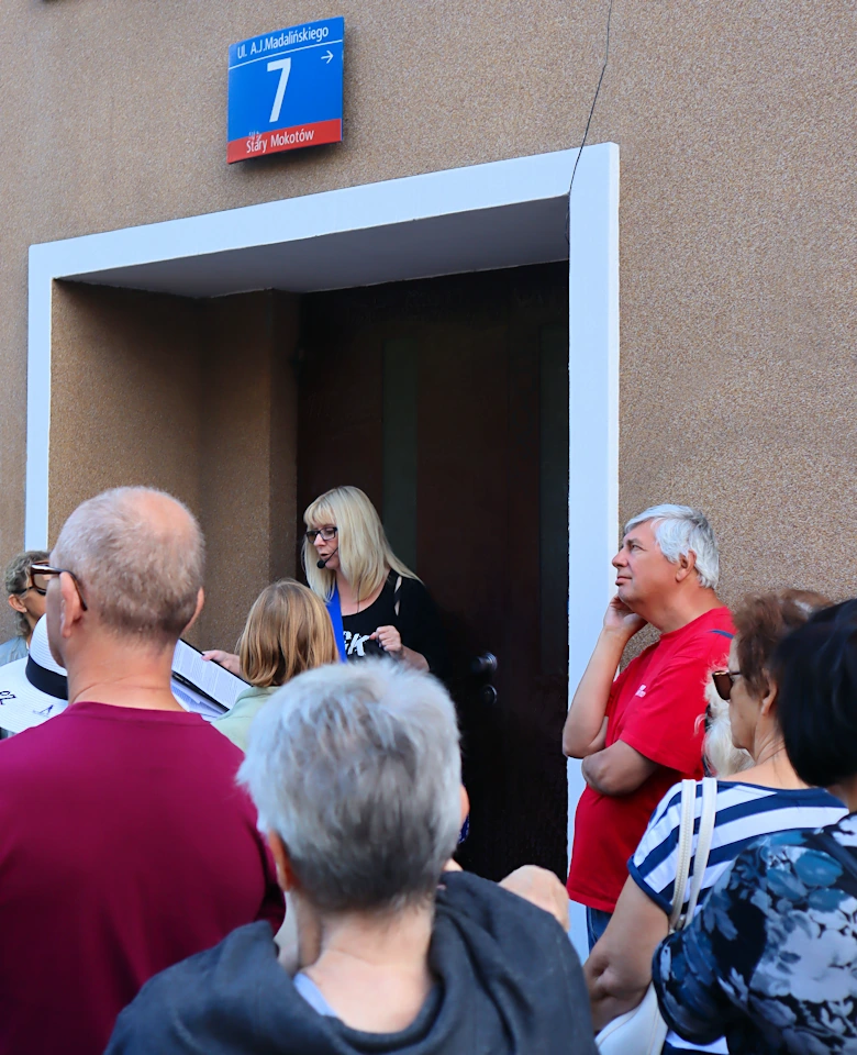 Grupa osób w letnich strojach stoi przed wejściem do budynku. W wejściu stoi przewodniczka, o czymś opowiadając.
