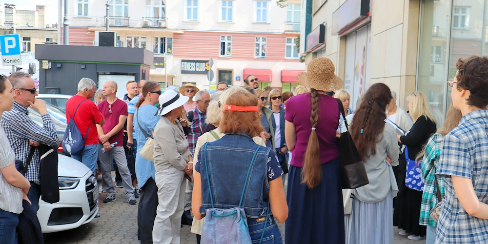 Grupa ludzi stoi przed budynkiem z prawej. Przewodniczka opowiada o czymś. Z lewej widać parkujące samochody. W tle widać poprzecznie stojący kilkupiętrowy budynek.