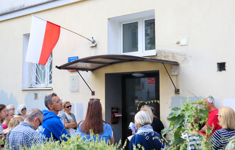 Grupa osób w letnich strojach stoi przed wejściem do budynku w jasnym tynku. W wejściu stoi przewodniczka, o czymś opowiadając.