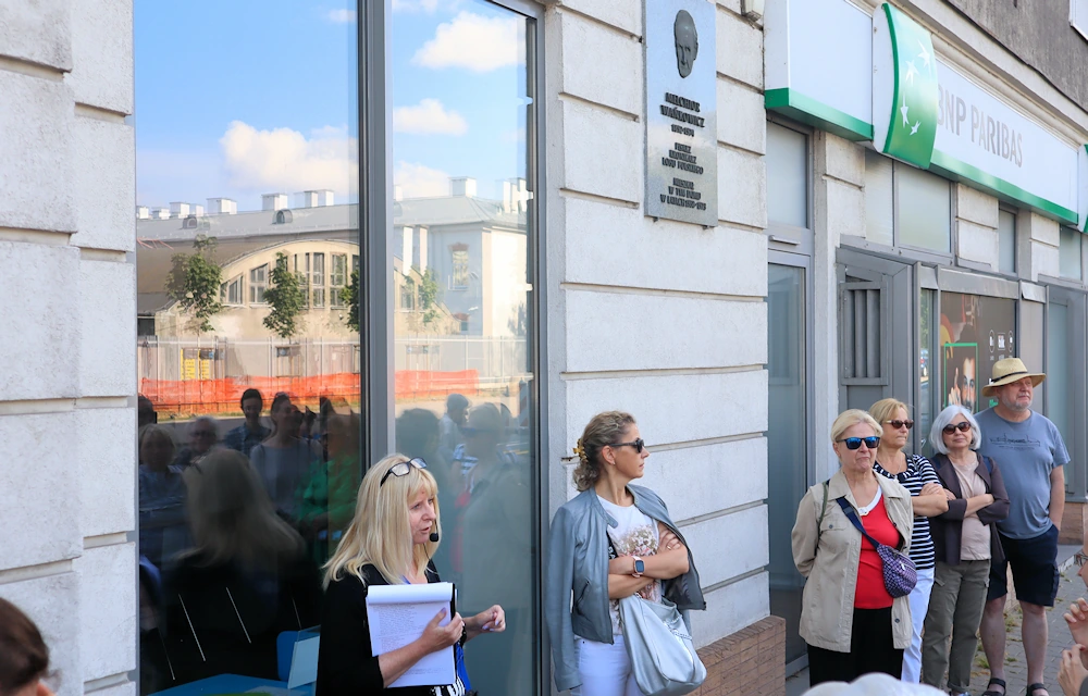 Grupa ludzi stoi przed bramą budynku, na którym powieszono tablicę pamiątkową z Wańkowiczem. To jasno otynkowana fasada i bardzo duże witryny na parterze.