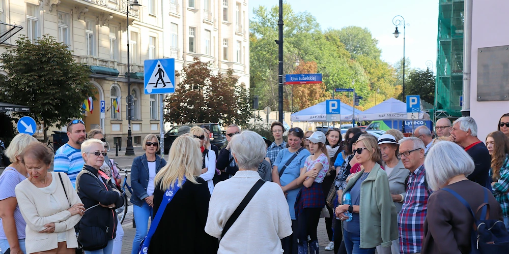 Grupa osób w letnich strojach stoi na chodniku przy rozległej ulicy. Z lewej widać fasadę jasnożółtego budynku przedwojennego. W tle po środku widać drzewa liściaste. Po prawej fragment budynku pomalowanego na fioletowo.