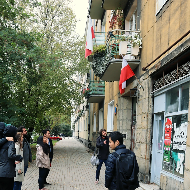Z prawej niska kamienica ze sklepem na dole, balkonami i polskimi flagami. Wzdłuż chodnika stoją uczestnicy spaceru słuchający przewodnika.
